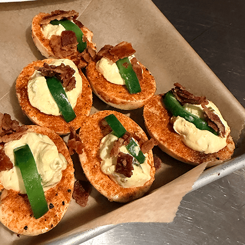 Crostini halves topped with whipped cream cheese, a strip of green garnish, and crumbled bacon pieces, arranged on parchment