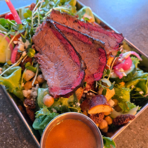 Slices of medium-rare beef resting on a greens salad with chickpeas, croutons, and microgreens, accompanied by a small cup of dressing.