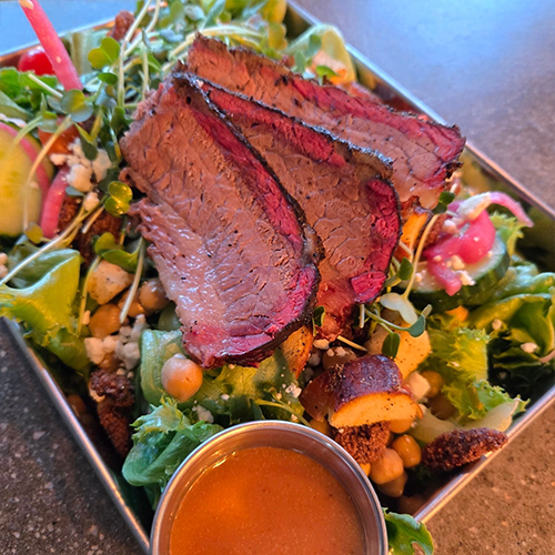 Slices of medium-rare beef resting on a greens salad with chickpeas, croutons, and microgreens, accompanied by a small cup of dressing.