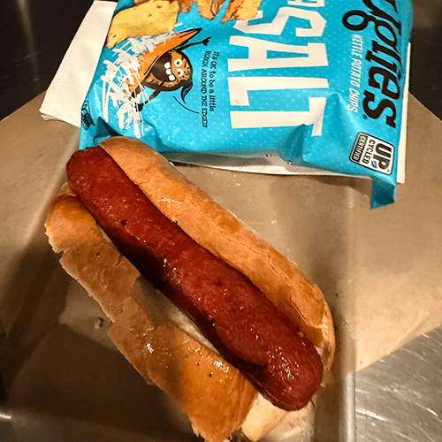 Hot dog in a soft bun on a stainless steel surface with a blue chip bag in the background pale lighting behind it.