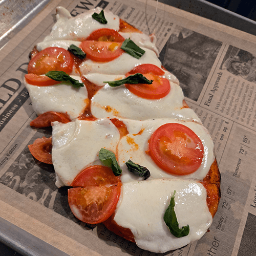 Flatbread pizza on a newspaper-lined tray topped with mozzarella, tomato slices, and fresh basil.