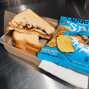 PB&J-style sandwich halves with jam filling on white bread, placed on brown paper next to a blue bag of potato chips on a metal counter