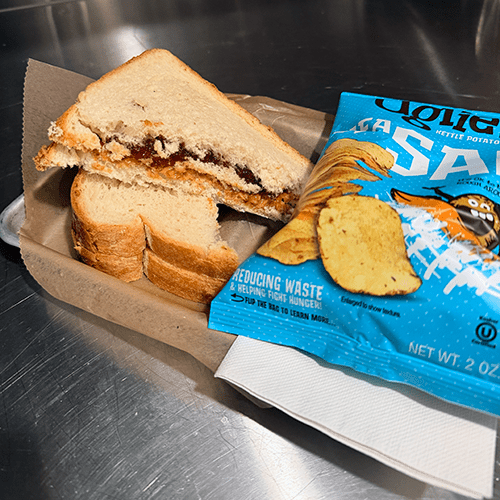 PB&J-style sandwich halves with jam filling on white bread, placed on brown paper next to a blue bag of potato chips on a metal counter