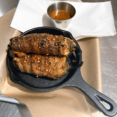 Two bacon-wrapped meat rolls in a cast-iron skillet, seasoned and crusty, with a small cup of dipping sauce in the background.