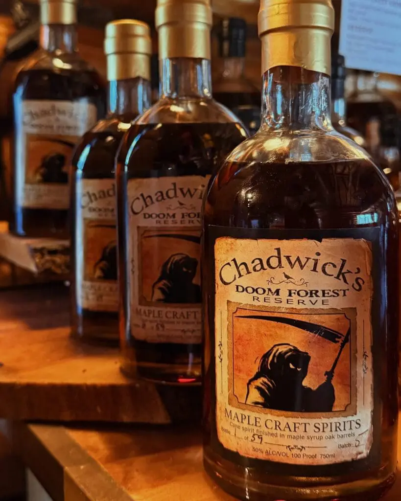 Close-up bottle of Chadwick's Maple Craft Spirits on a wooden shelf; label reads Doom Forest Reserve and shows a vintage drawing.