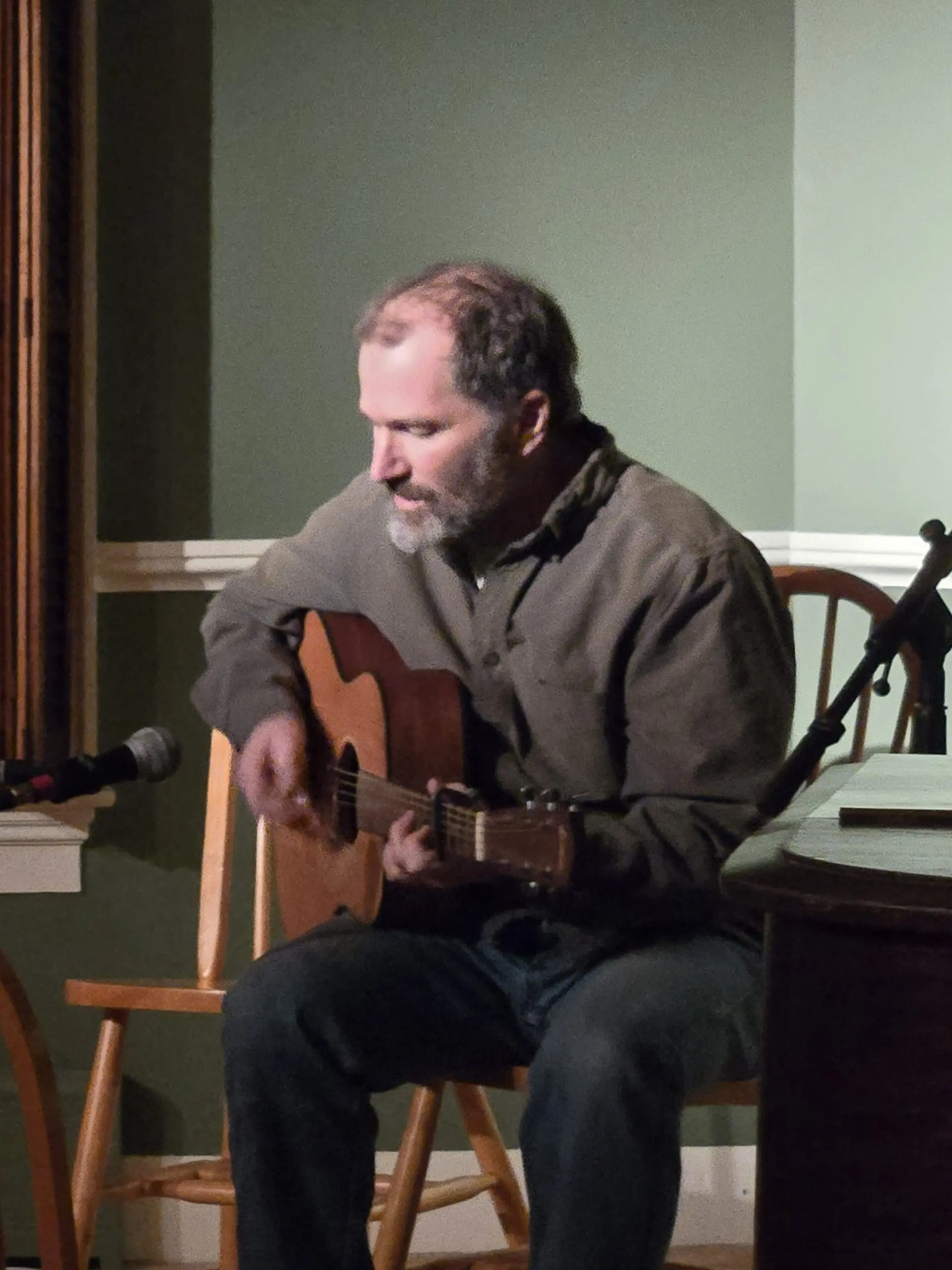 Man seated and playing an acoustic guitar, with a microphone nearby in a cozy room.