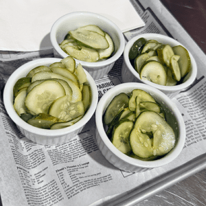 Four white ramekins of sliced cucumber pickles on a newspaper-lined tray.