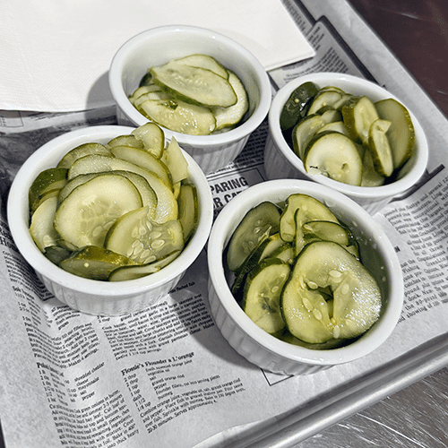 Four white ramekins of sliced cucumber pickles on a newspaper-lined tray.