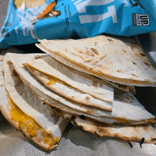 Stack of folded quesadilla wedges with melted cheese on a kitchen counter, near a blue bag of chips in the background.